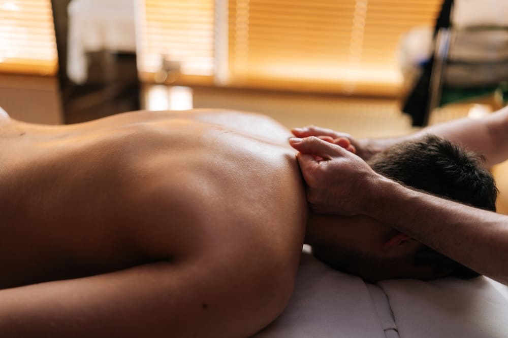 A man lies face down on a towel and receives a therapeutic massage on his neck